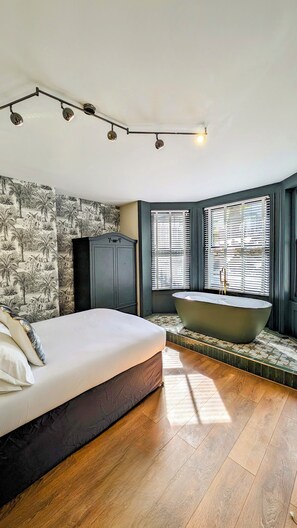 A bedroom with a large bed, a dark wardrobe, a freestanding bathtub, and a window with blinds. - INNit Bliss (Brighton)
