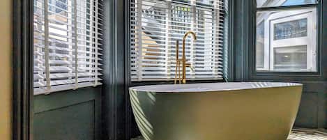 Un baño moderno con una bañera independiente, ventanas amplias con persianas y piso de baldosas.