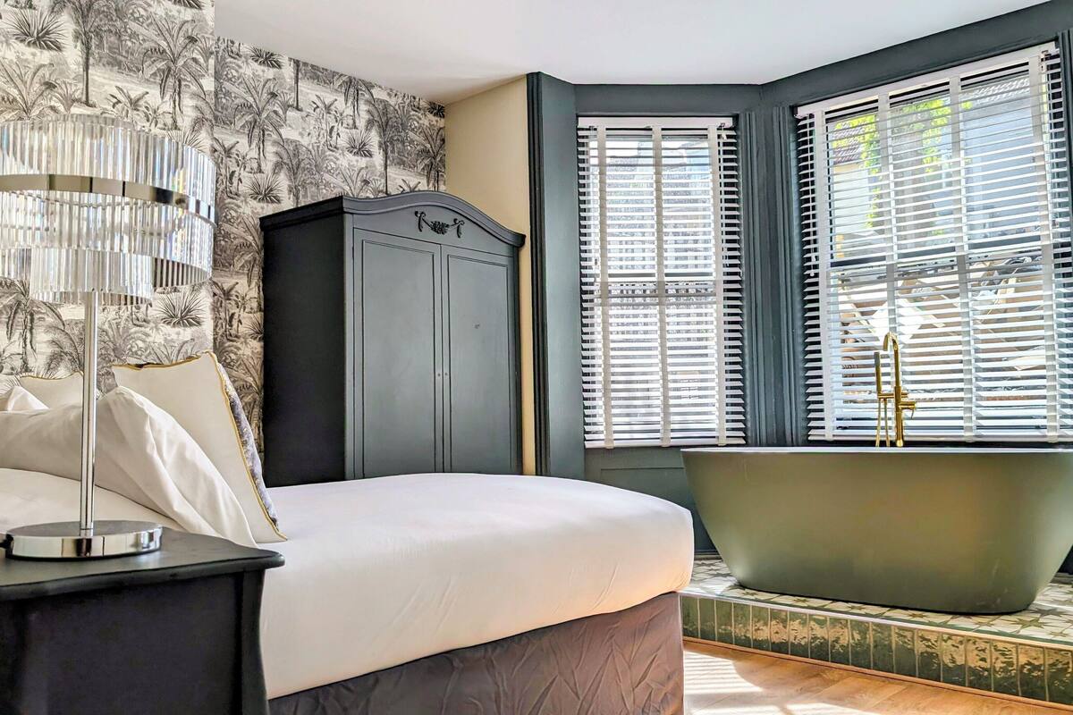 A bedroom with a bed, a nightstand, a lamp, a wardrobe, and a bathtub.