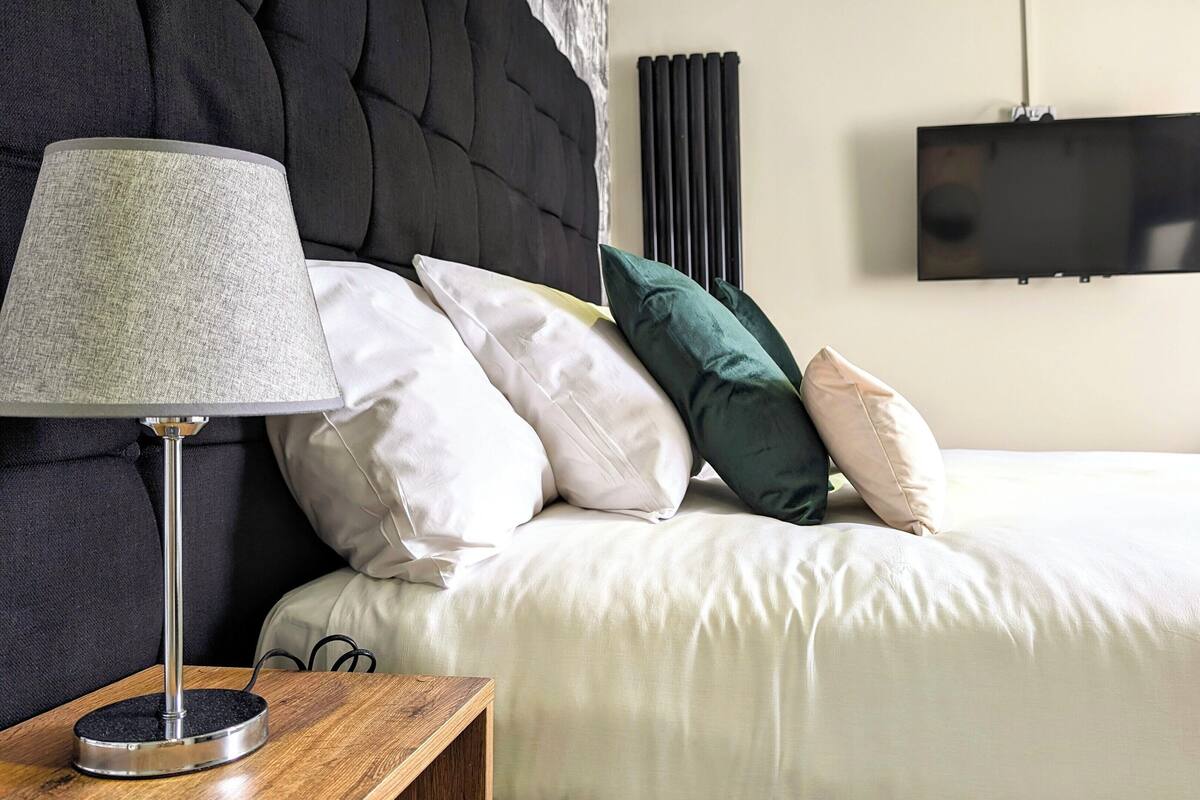 A neatly made bed with white bedding, a round table with a lamp, and a television on the wall.