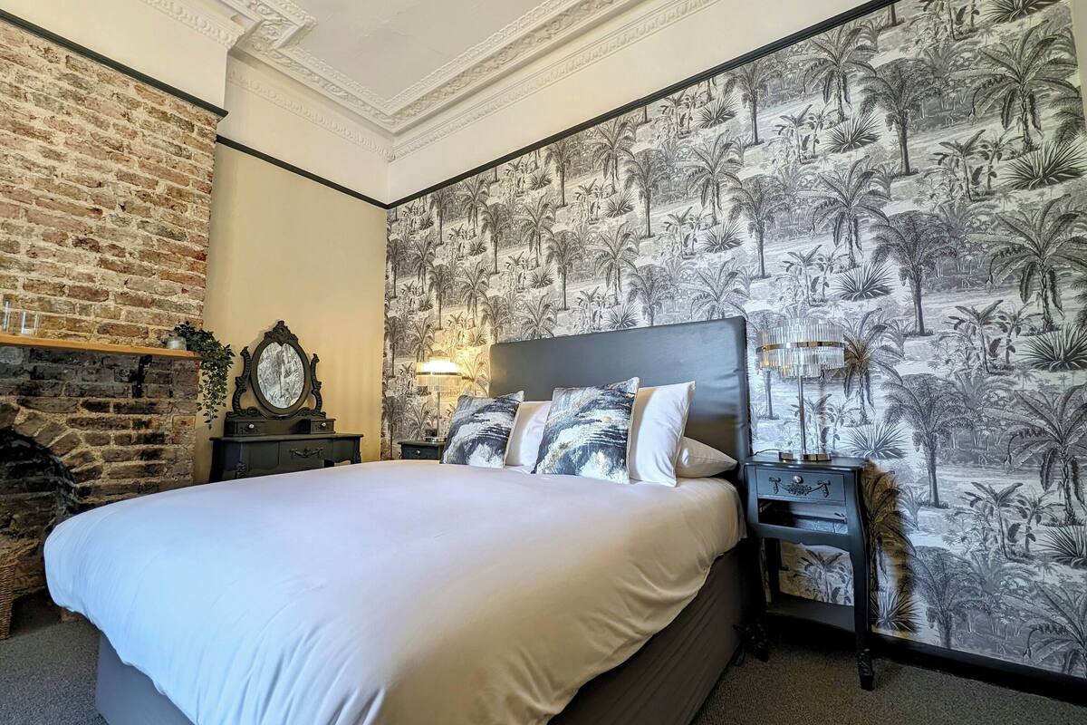 A bedroom with a brick wall, a bed with white linens, a nightstand with a lamp, and a wall with a tropical wallpaper.
