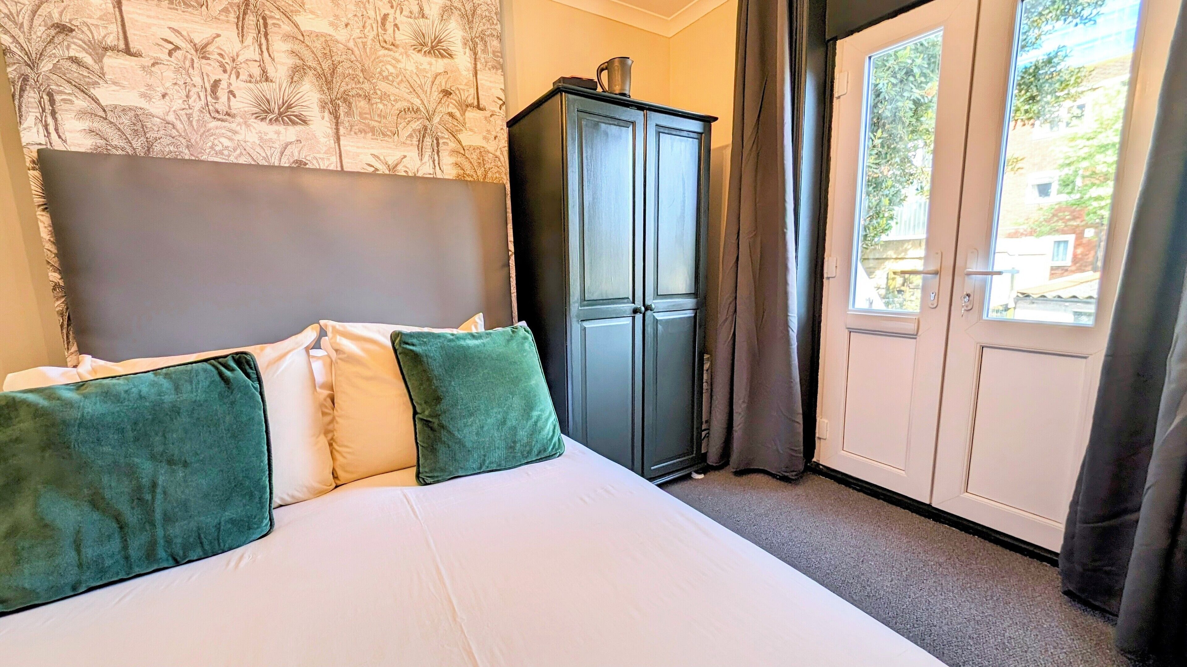 A bedroom with a bed, green pillows, a dark cabinet, and a view of the outside through a door.