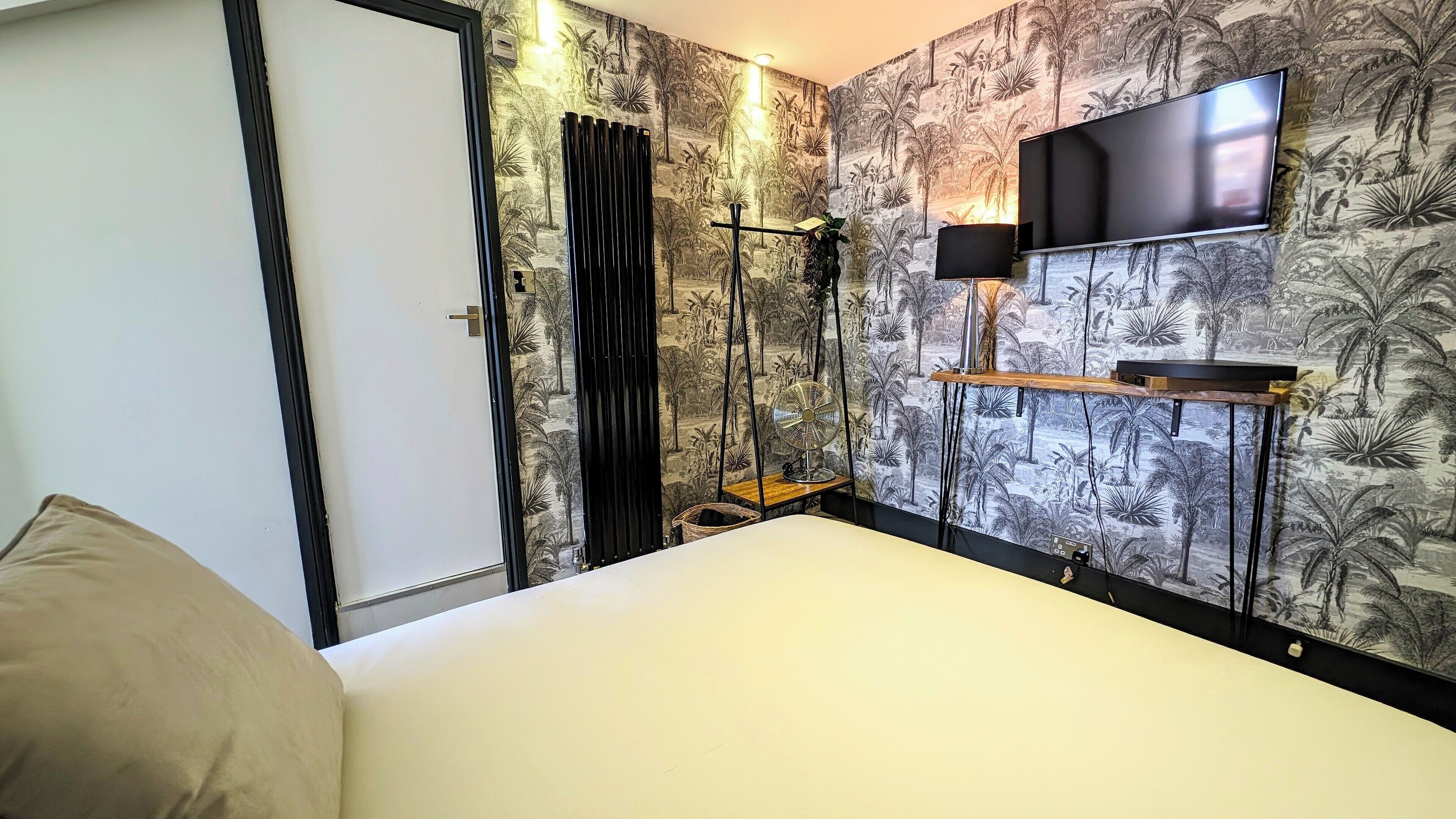 A hotel room with a bed, a TV mounted on the wall, a fan, and a shelf with a plant.