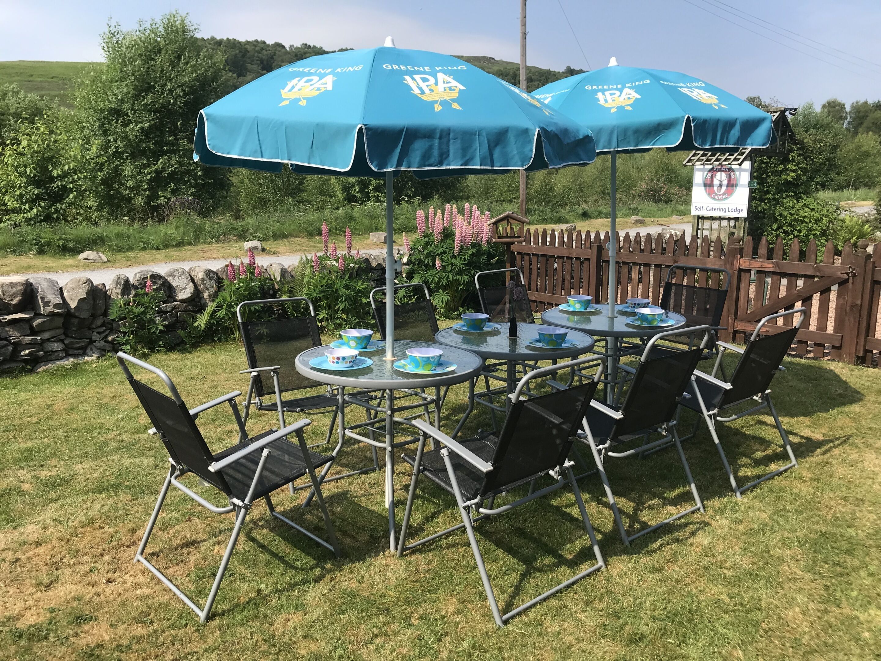 A self-catering lodge with outdoor seating under blue umbrellas, a stone wall, and a wooden fence.