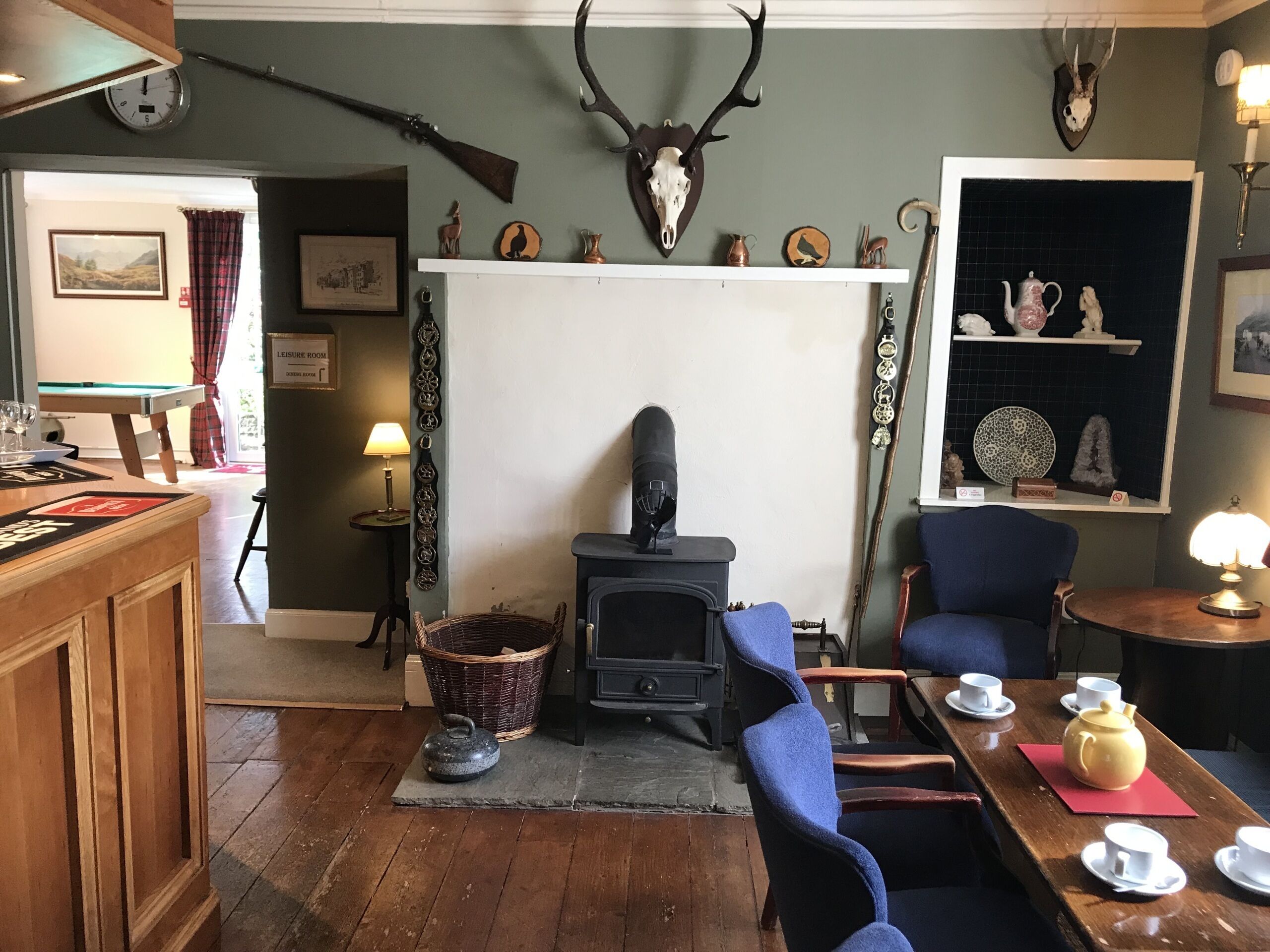 A cozy room with a fireplace, a rustic wooden table with cups and a teapot, and a wall adorned with antlers and a clock.