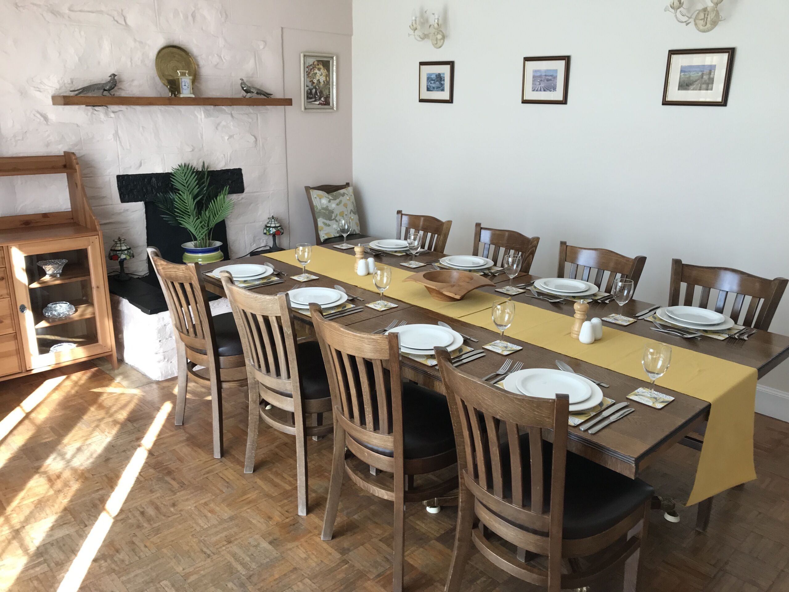 A dining area with a long table set for 10, surrounded by wooden chairs. There are wall-mounted shelves with decorative items and framed pictures.