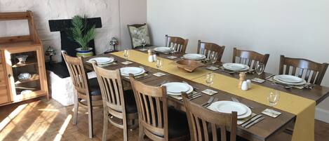 A dining area with a long table set for 10, surrounded by wooden chairs. There are wall-mounted shelves with decorative items and framed pictures.