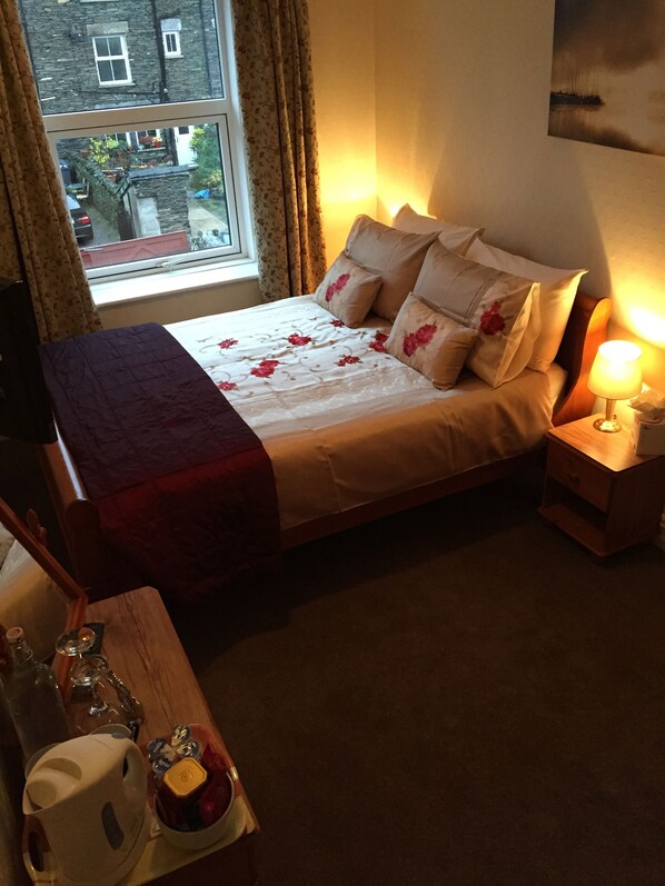 A neatly made bed with floral bedding, a bedside lamp, and a view of the outside through a window.