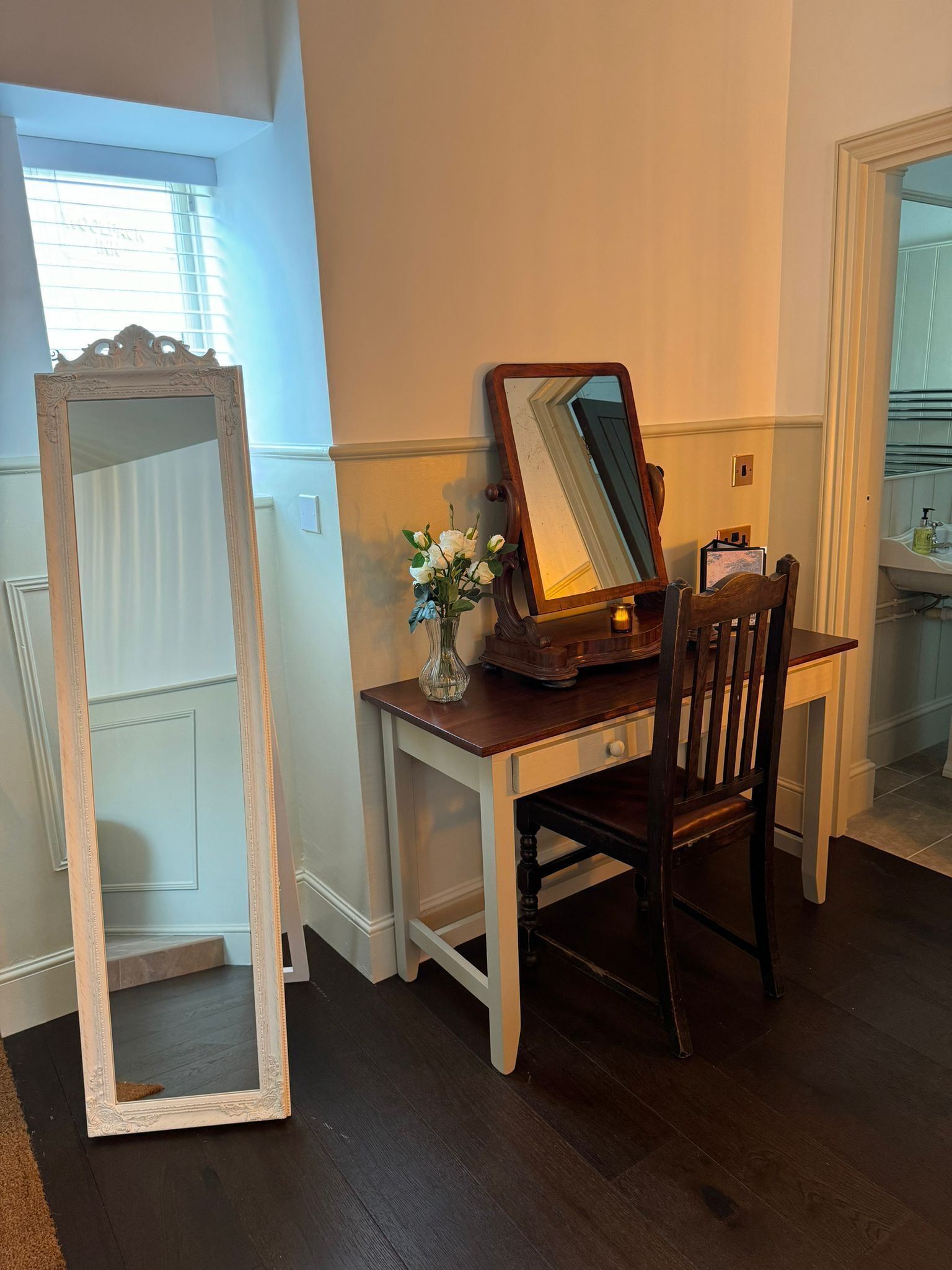 A room with a wooden desk, a mirror, and a chair. There is a vase with flowers on the desk.
