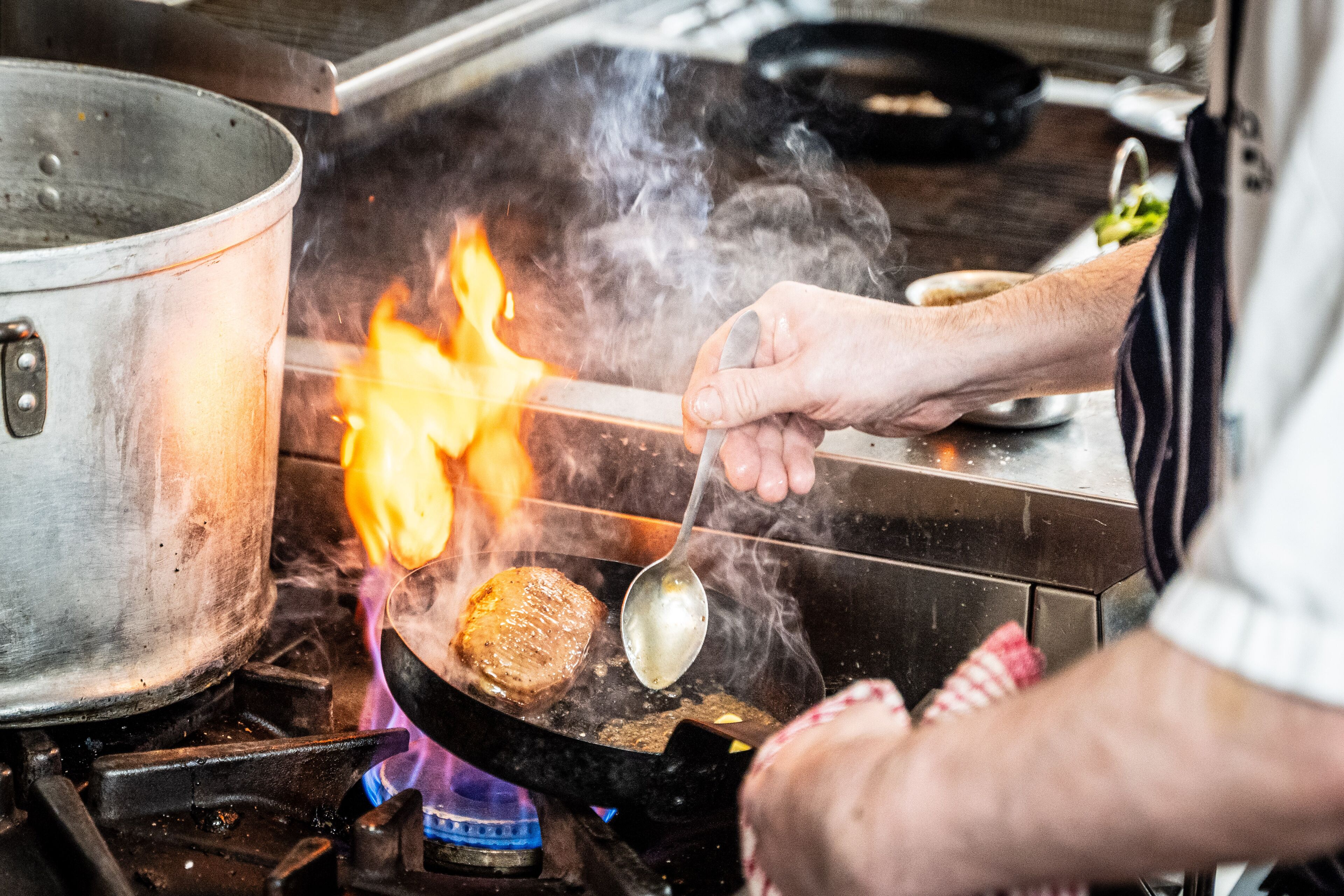 A cooking scene with a pan on a stove, a pot, and a person cooking a steak.