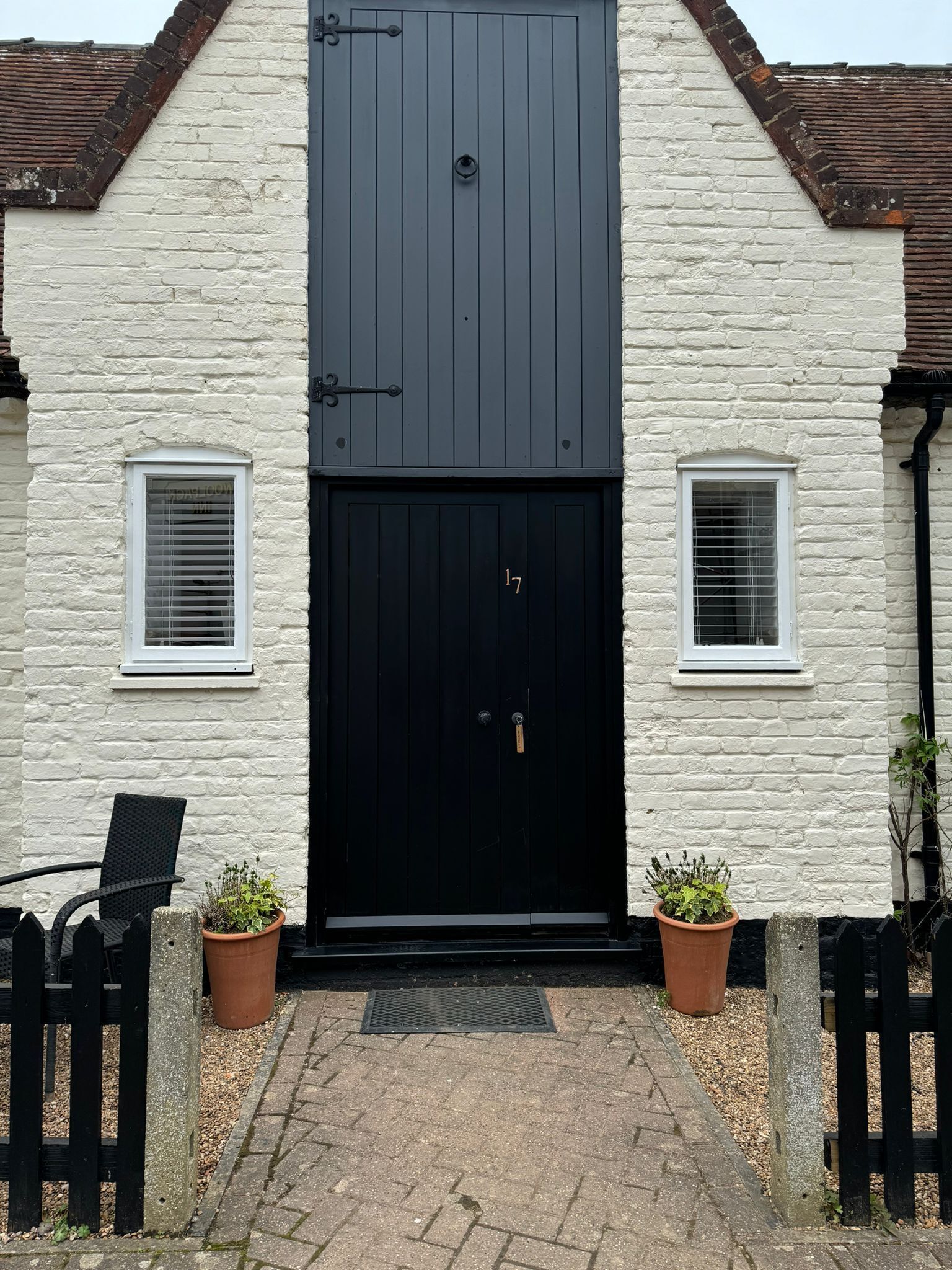 A white building with a black door and two windows. There are potted plants on either side of the door.