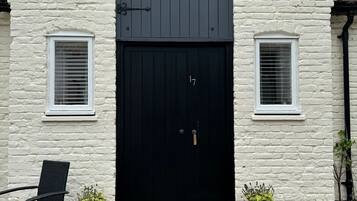 A white building with a black door and two windows. There are potted plants on either side of the door.