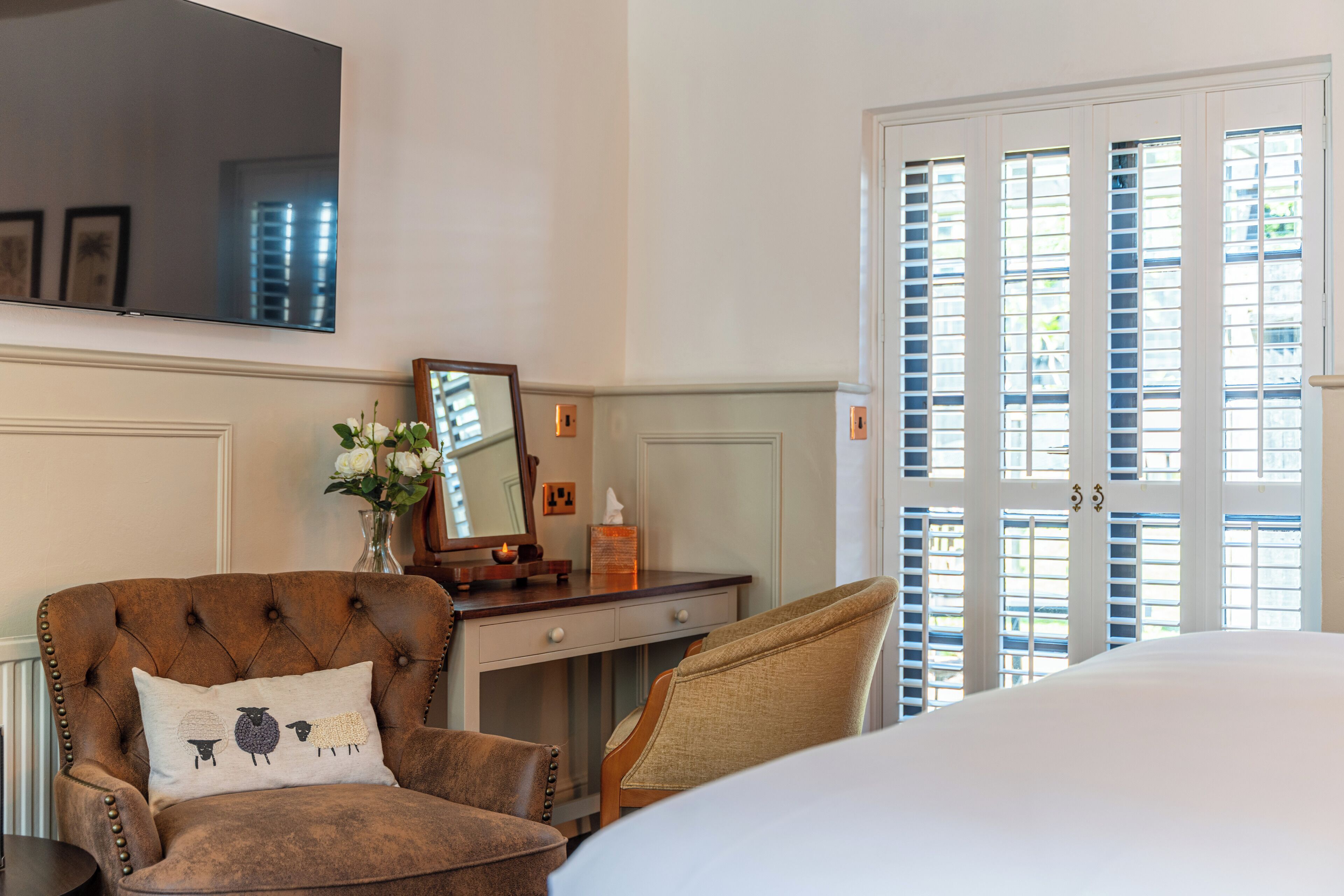 Habitación de hotel con una cama grande, un rincón de descanso con una silla y una mesita, un aparador, un espejo y una ventana con persianas.