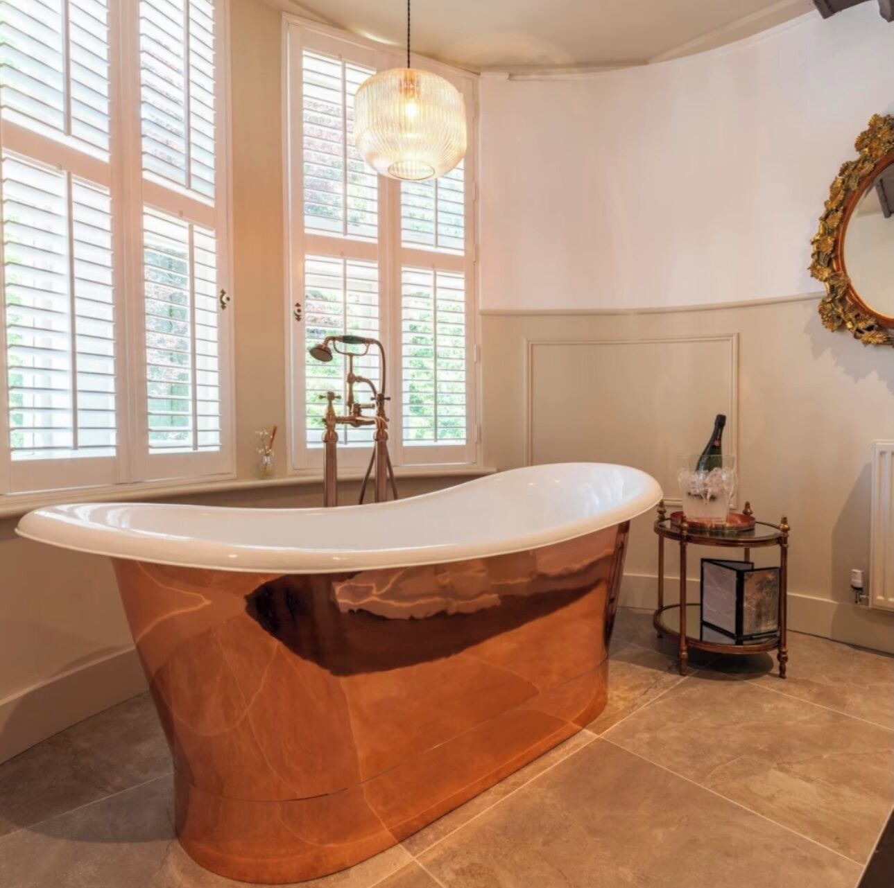 A spacious bathroom with a freestanding bathtub, large windows, a chandelier, and a mirror with a decorative frame.