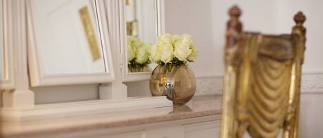 A white vanity with a gold-framed mirror, a vase with white flowers, and a gold-framed chair.
