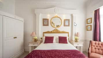 Una habitación de hotel con una cama grande, cabecera con marco dorado, dos mesitas de noche con lámparas y un espejo grande sobre la cama.