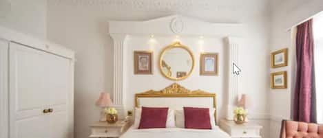 Une chambre d’hôtel avec un grand lit, une tête de lit ornée d’un cadre doré, deux tables de chevet avec des lampes et un grand miroir au-dessus du lit.