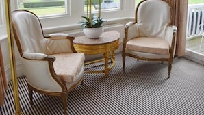 A room with two white upholstered chairs, a small round table with a white vase and plant, and a lamp. Large windows with white frames and curtains are visible. - The Lookout Rye (Rye)