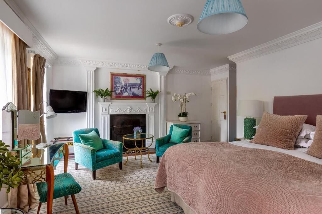 Una camera d'albergo con un letto grande, due poltrone color petrolio, un tavolino rotondo, un caminetto e un televisore appeso al muro.