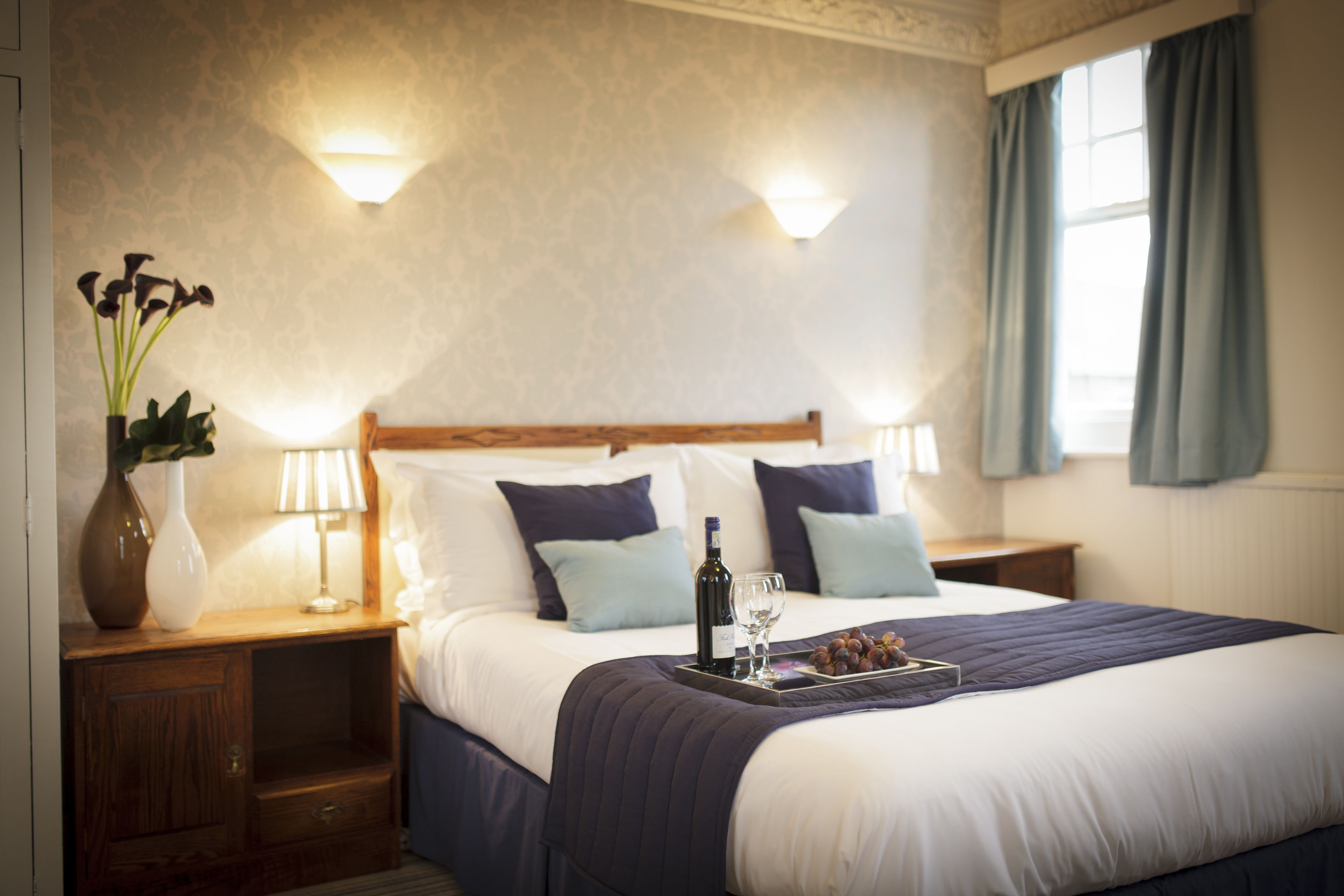 A neatly made bed with a tray of grapes and a bottle, two bedside lamps, and a vase with flowers on a wooden nightstand.