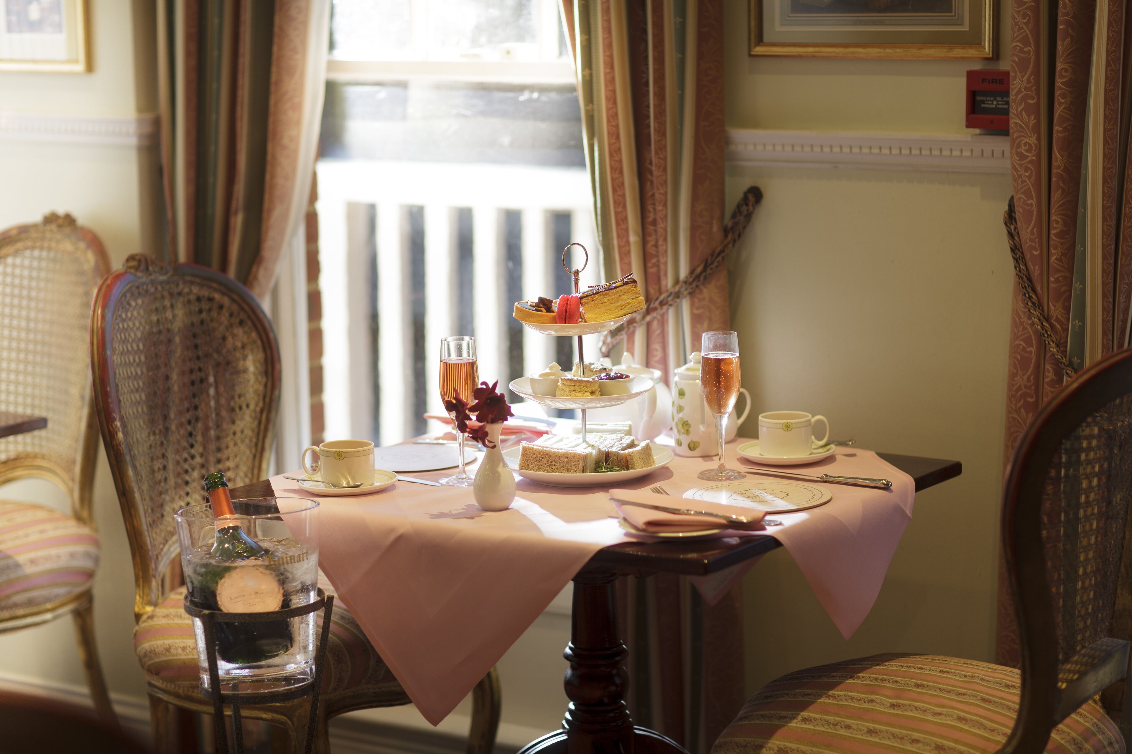 A dining table set with a tiered cake, sandwiches, and drinks, surrounded by upholstered chairs.