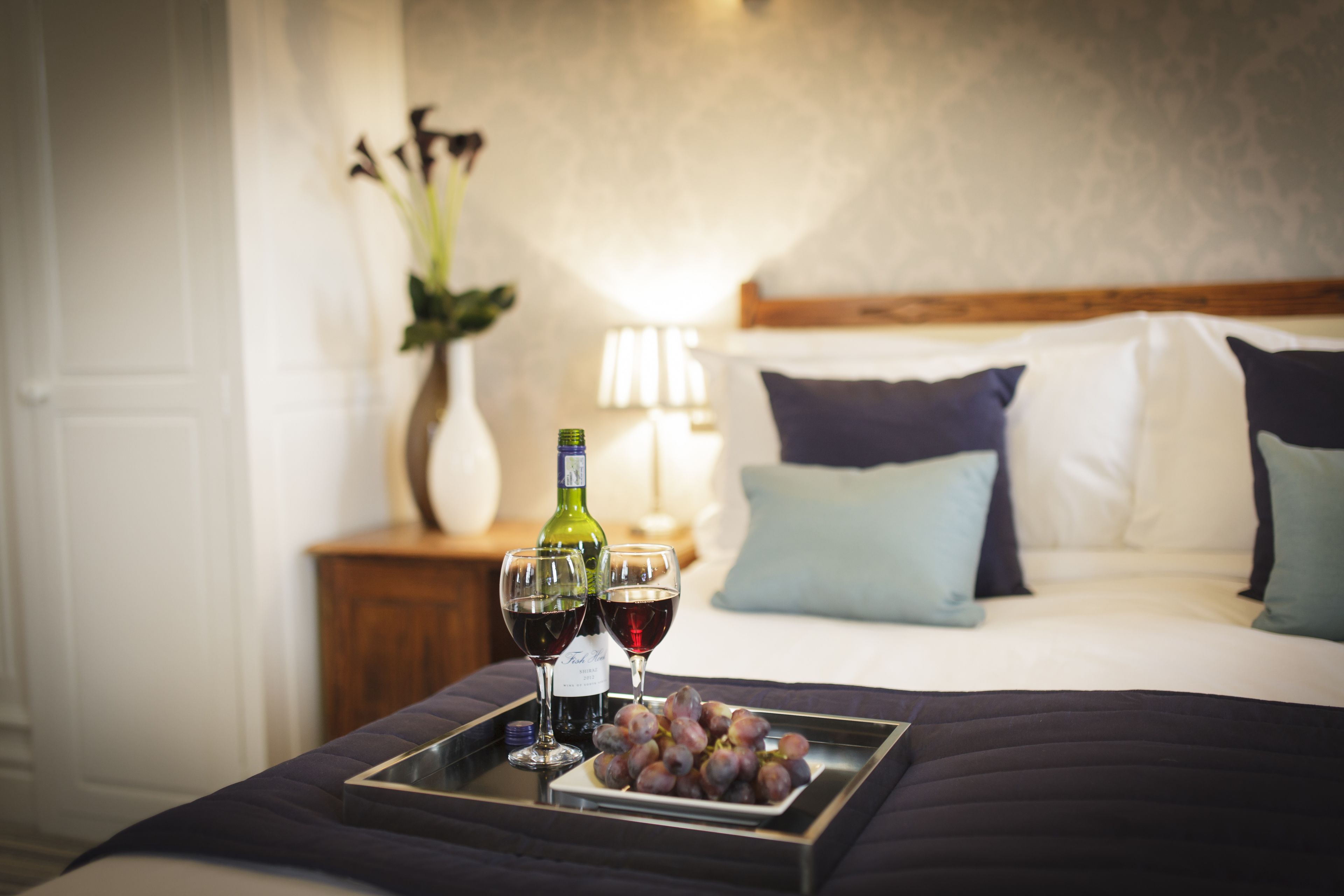 Una cama bien hecha con una colcha azul oscuro, una bandeja con dos copas de vino y un plato de uvas, y una botella verde de vino sobre una mesita de noche.