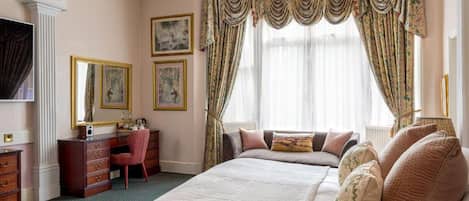 A bedroom with a large bed, a desk, and a chair. There are curtains, a chandelier, and framed pictures on the wall.
