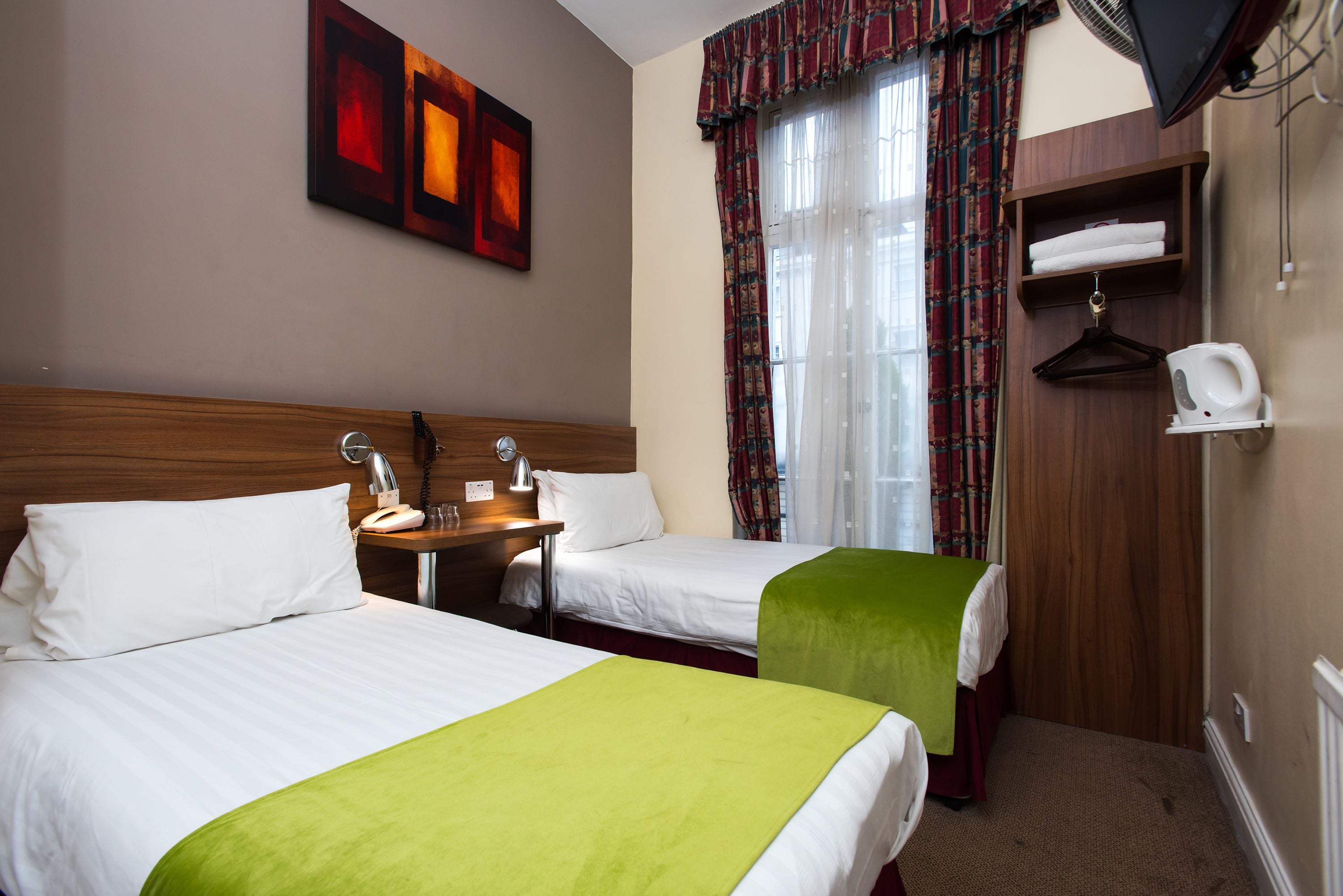 A hotel room with two beds, a bedside table with a lamp, a TV mounted on the wall, and a window with curtains.