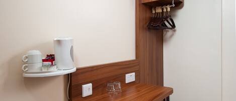 A compact hotel room with a wooden desk, a red chair, a television mounted on the wall, a small shelf with cups and a coffee maker, and a coat rack with hangers.