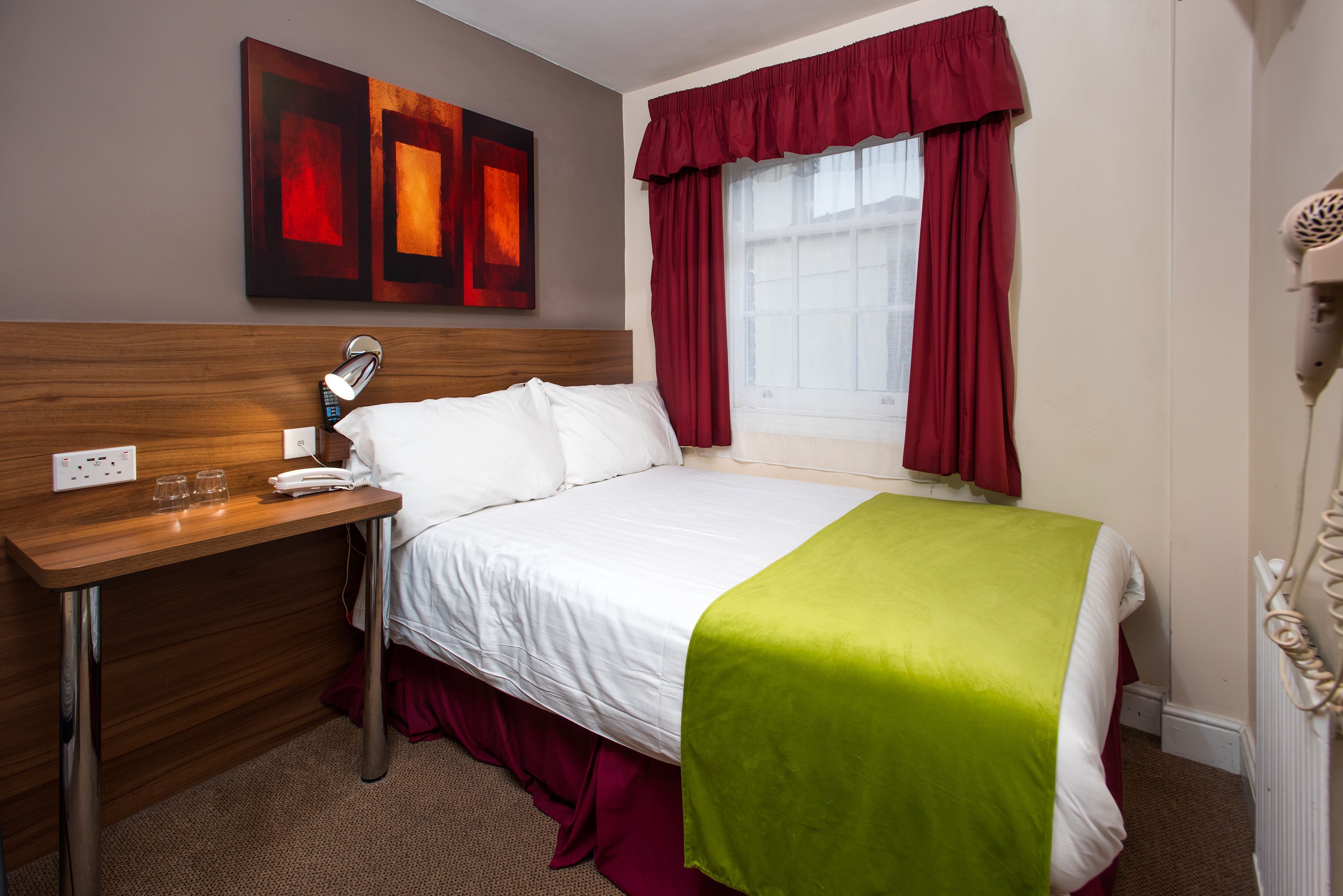 Une chambre d’hôtel avec une tête de lit en bois, un lit avec une literie blanche et un plaid vert, un bureau avec une lampe et une fenêtre avec des rideaux rouges.