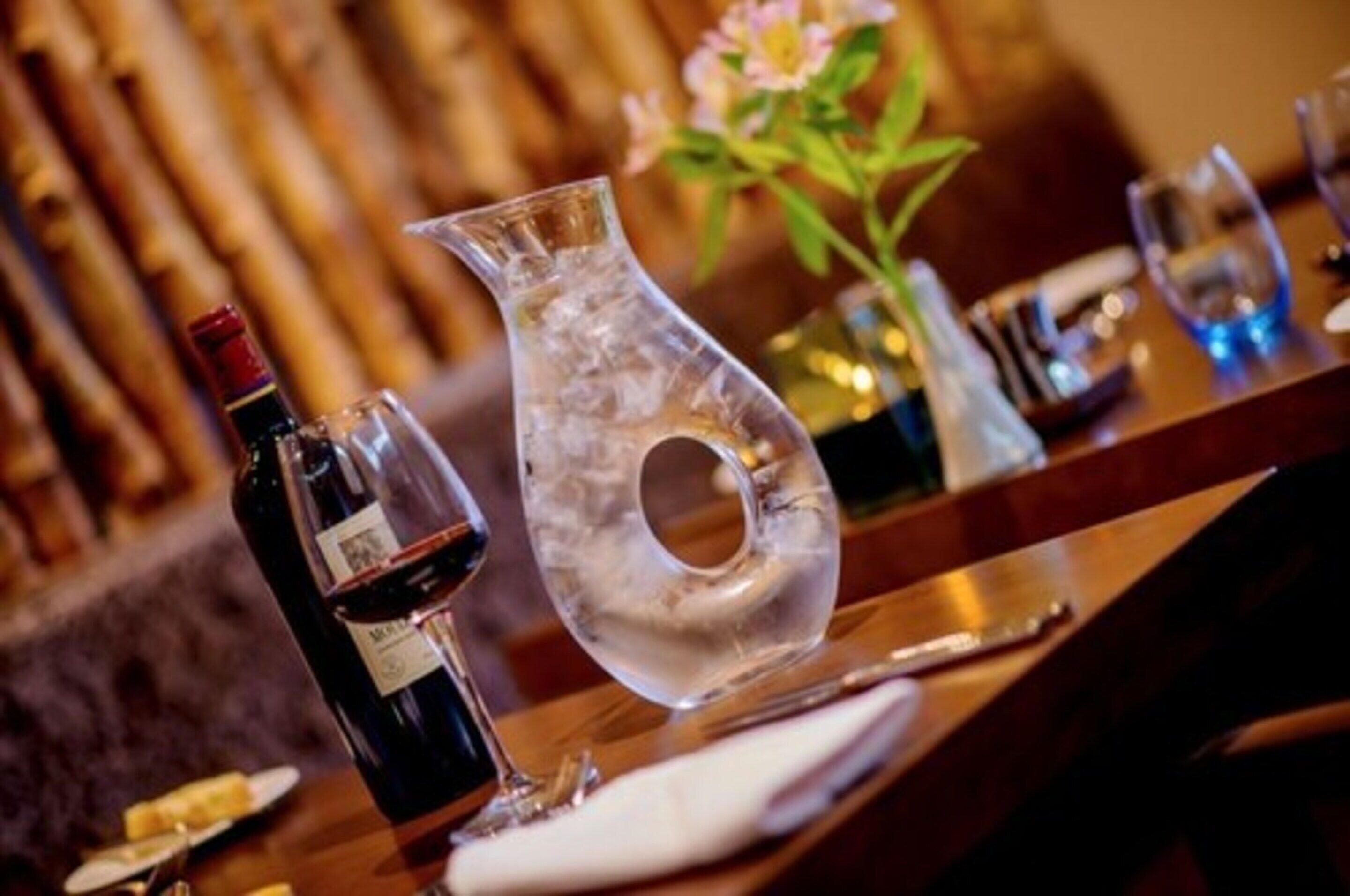 Una mesa de comedor con una botella de vino, una jarra de vidrio transparente y un florero con flores.