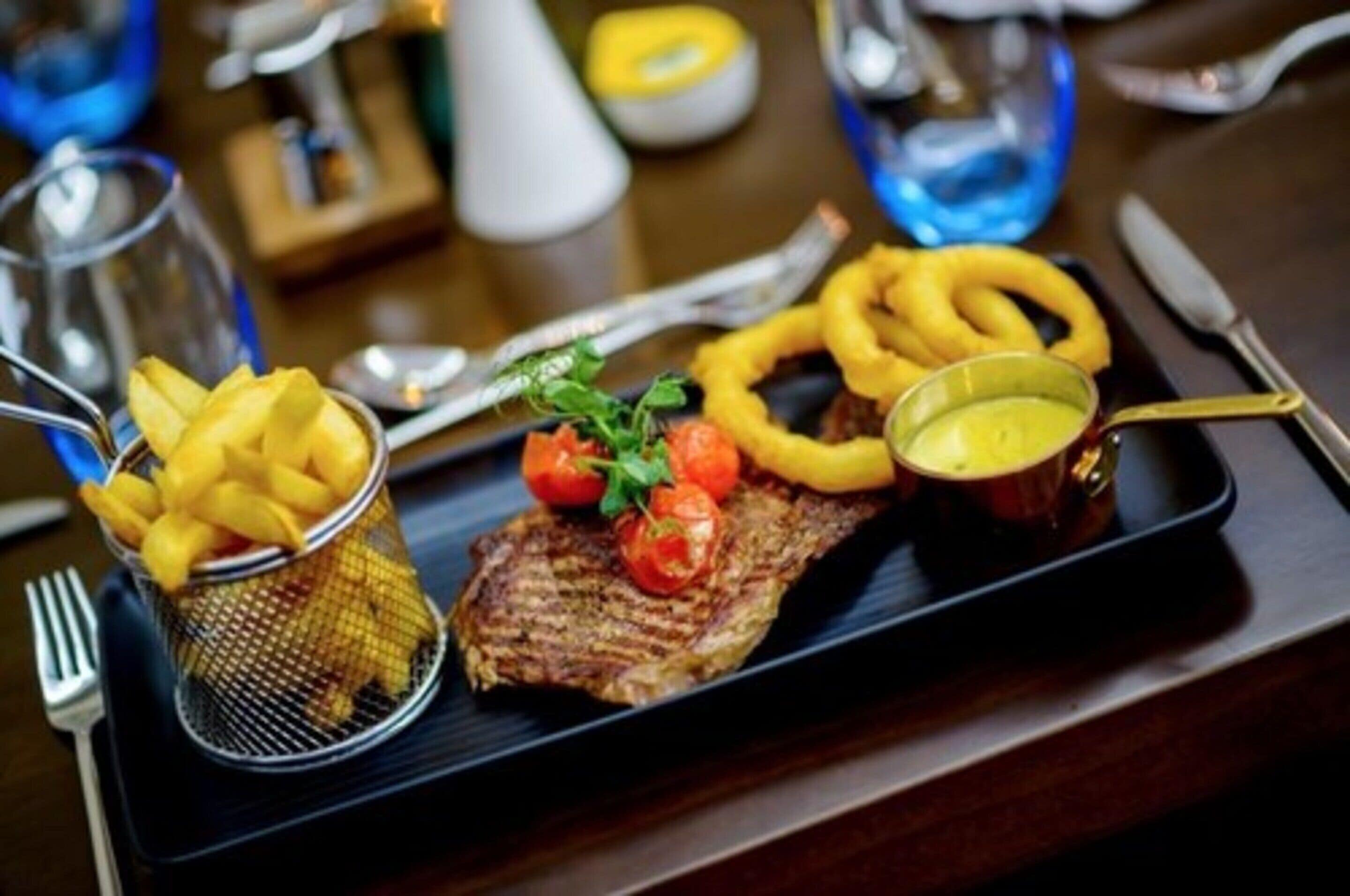 Un plato de carne con tomates y cebollas a la parrilla, acompañado de papas fritas y un recipiente pequeño de salsa, sobre una bandeja negra.
