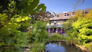 A garden with a pond, surrounded by lush greenery and a building with outdoor seating. - Le Friquet Country Hotel (Castel)