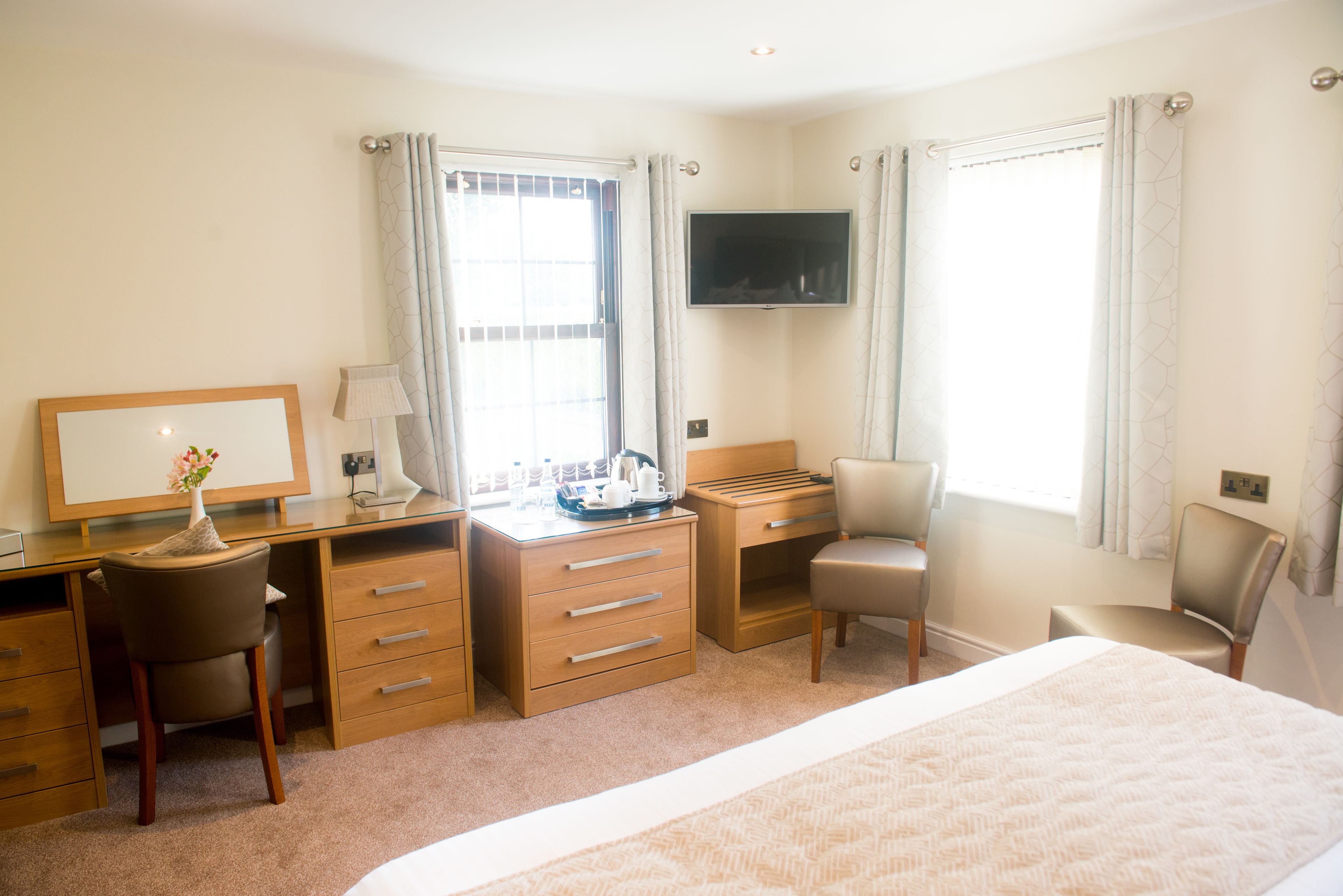 A hotel room with a bed, a desk with a chair, a TV mounted on the wall, and a window with curtains.