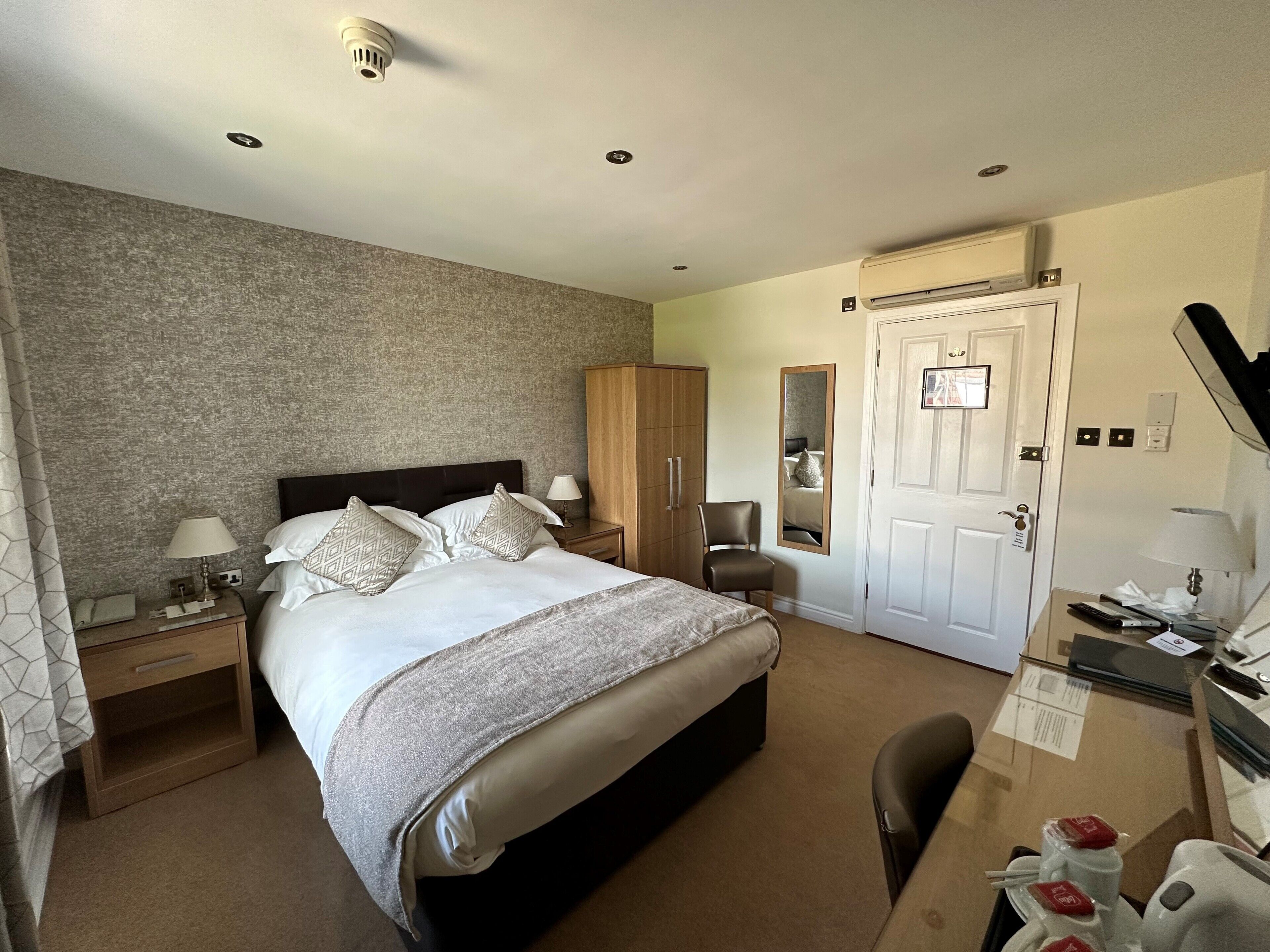 A hotel room with a large bed, a wardrobe, a mirror, a desk with a chair, and a door.