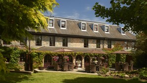 A stone building with a sloped roof and multiple windows. There are outdoor seating areas with umbrellas and potted plants. - Le Friquet Country Hotel (Castel)