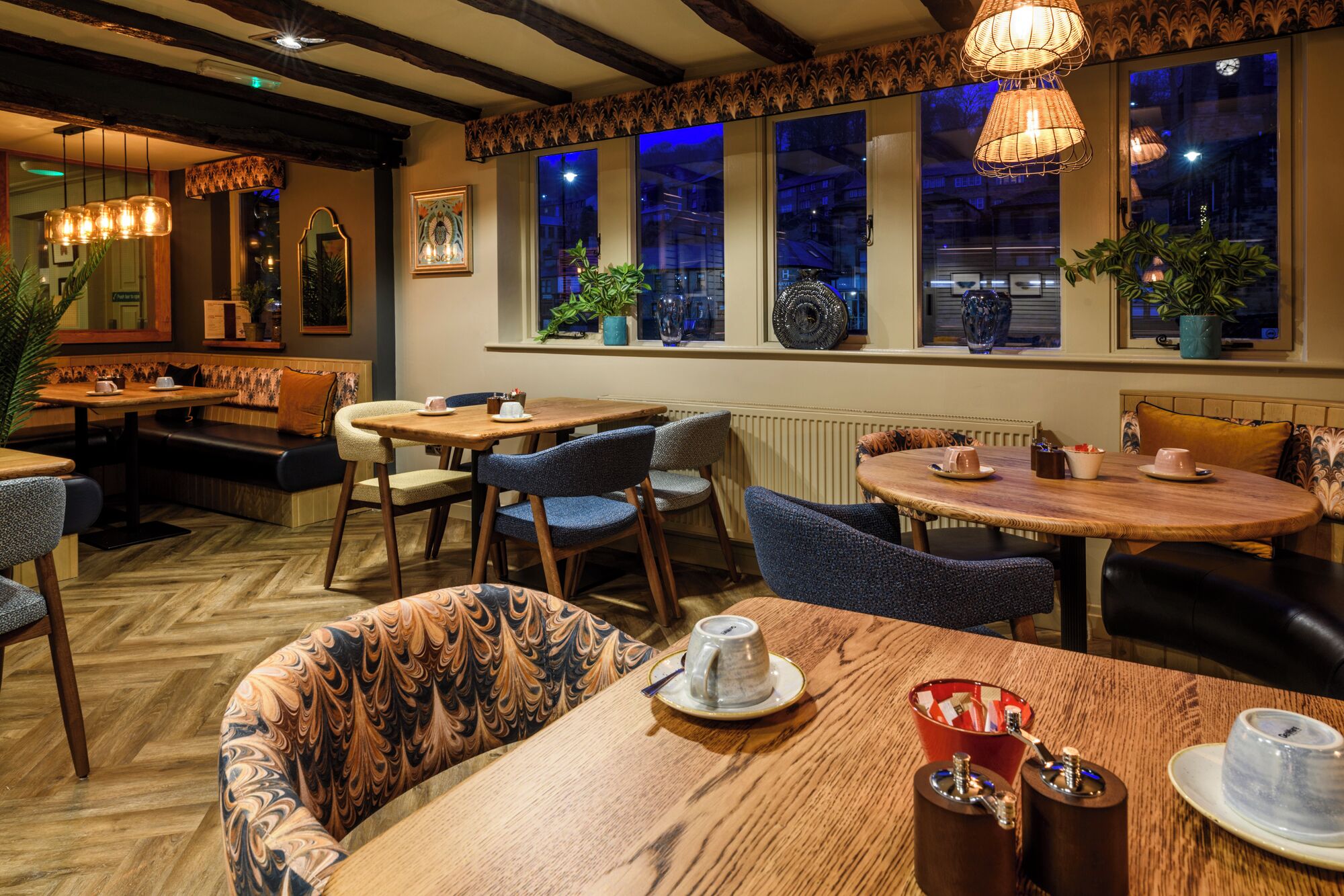A cozy restaurant with wooden tables and chairs, large windows with city views, and hanging wicker lamps.