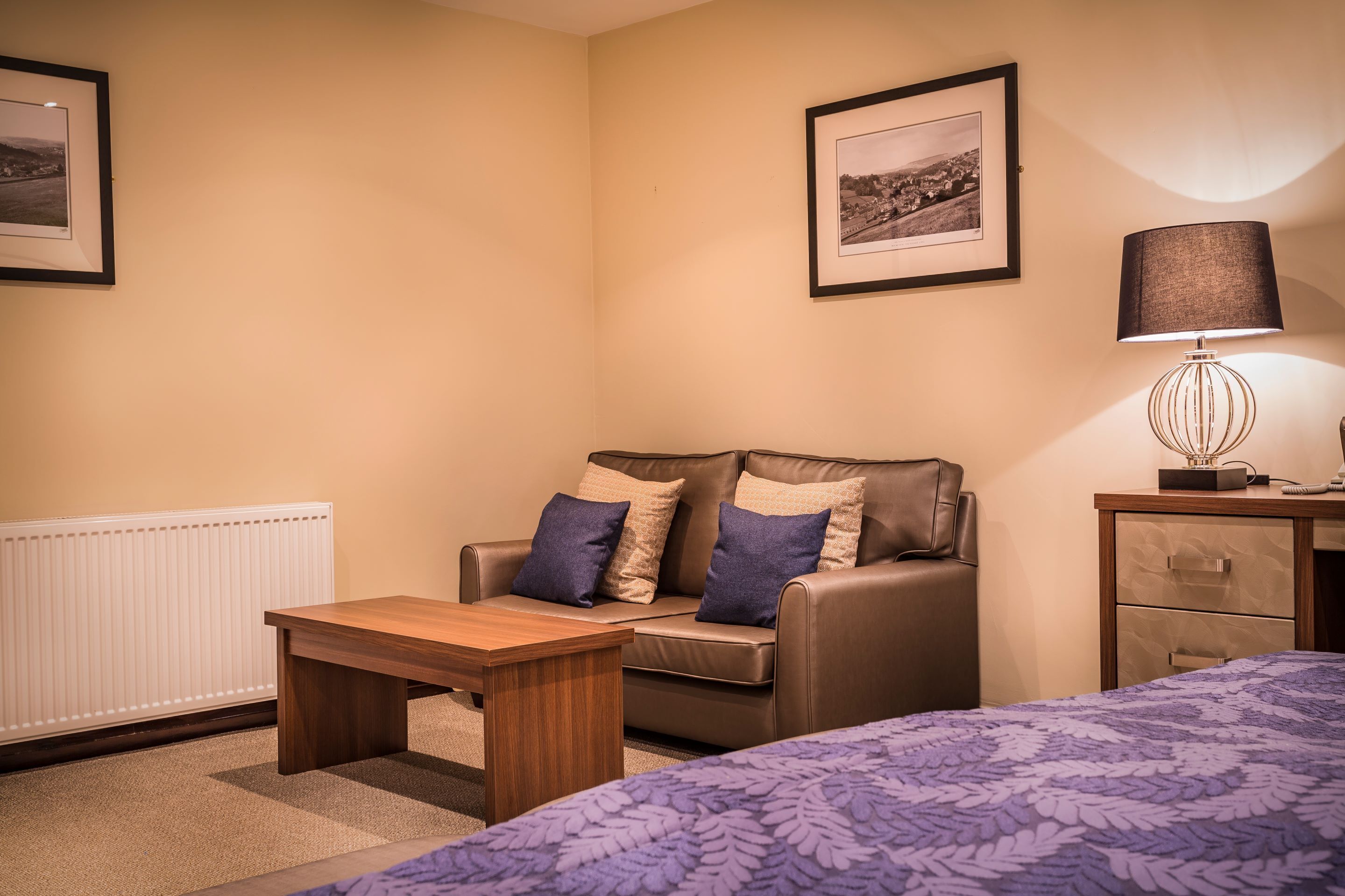 A hotel room with a bed, a wooden table, a sofa with cushions, a lamp, a framed picture on the wall, and a radiator.