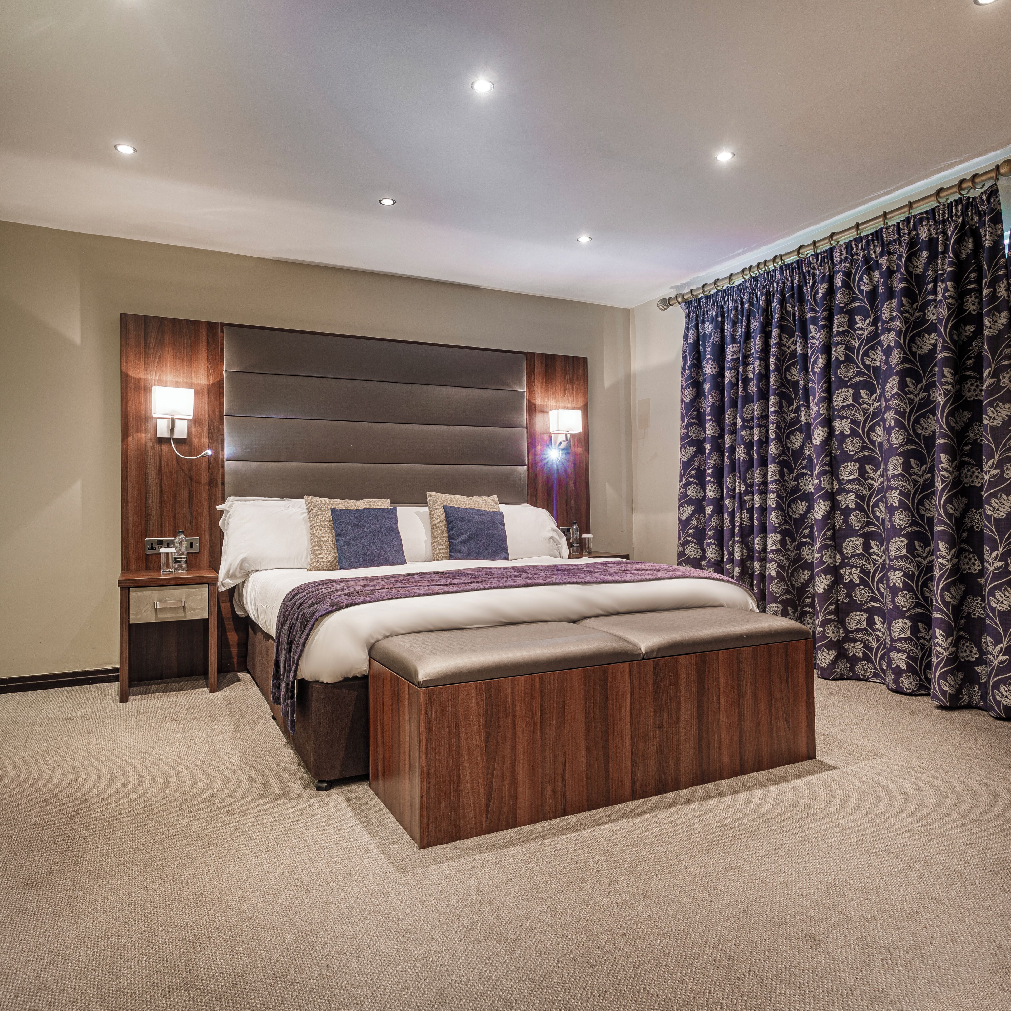 Un dormitorio de hotel moderno con una cama grande, cabecera de madera, lámparas de noche y cortina floral oscura.