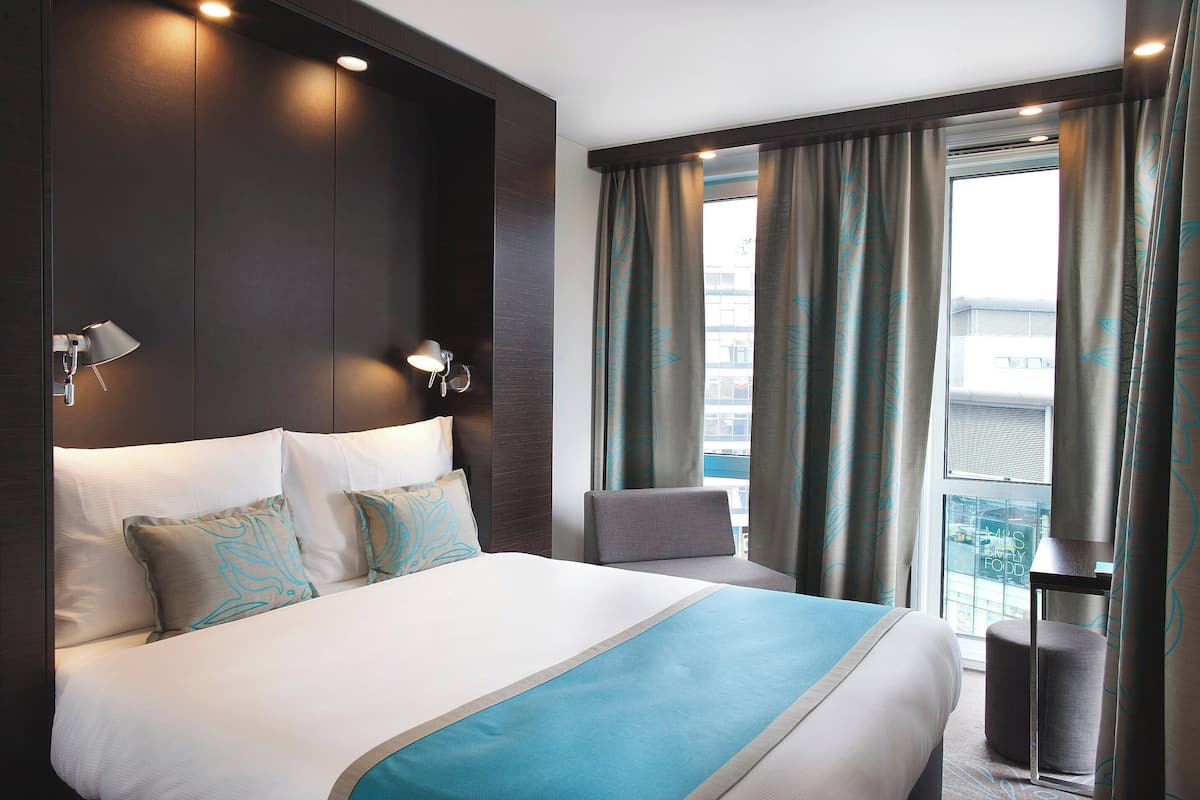 A hotel room with a large bed, a chair, a small table, and a window with curtains.
