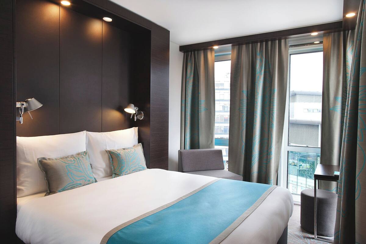 A hotel room with a large bed, a chair, a small table, and a window with curtains.