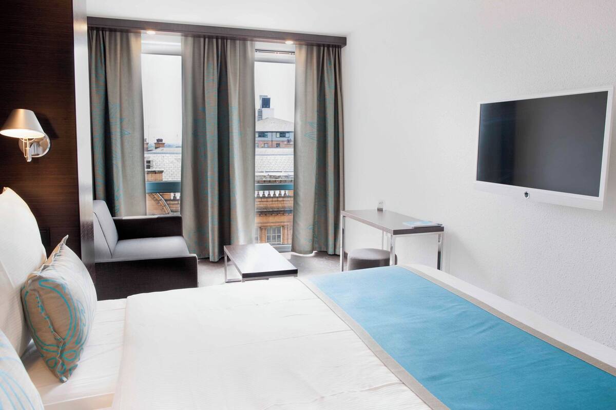 A hotel room with a bed, a desk, a chair, a sofa, a TV, and a window with curtains.
