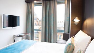 Habitación de hotel con cama, televisión montada en la pared, mesita de noche, lámpara y vistas a la ciudad a través de amplias ventanas.