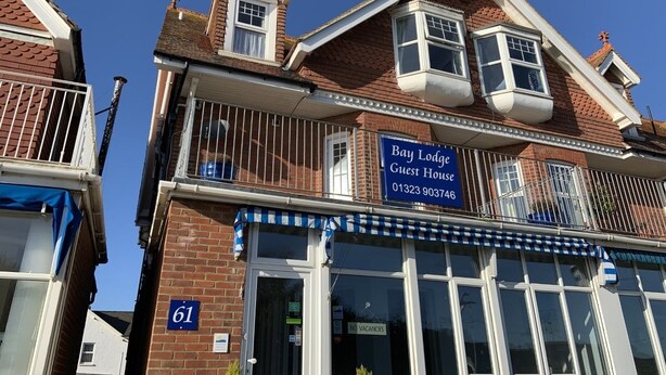 Ein zweistöckiges Gebäude mit einer blau-weißen Markise, einem Schild mit der Aufschrift "Bay Lodge Guest House" und der Hausnummer 61 an der Fassade.