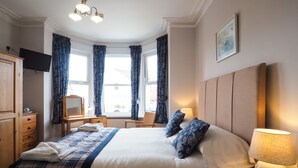 Ein Hotelzimmer mit einem großen Bett, einem Schreibtisch mit Lampe, einem Sessel, einem Fernseher und einem Fenster mit Vorhängen.