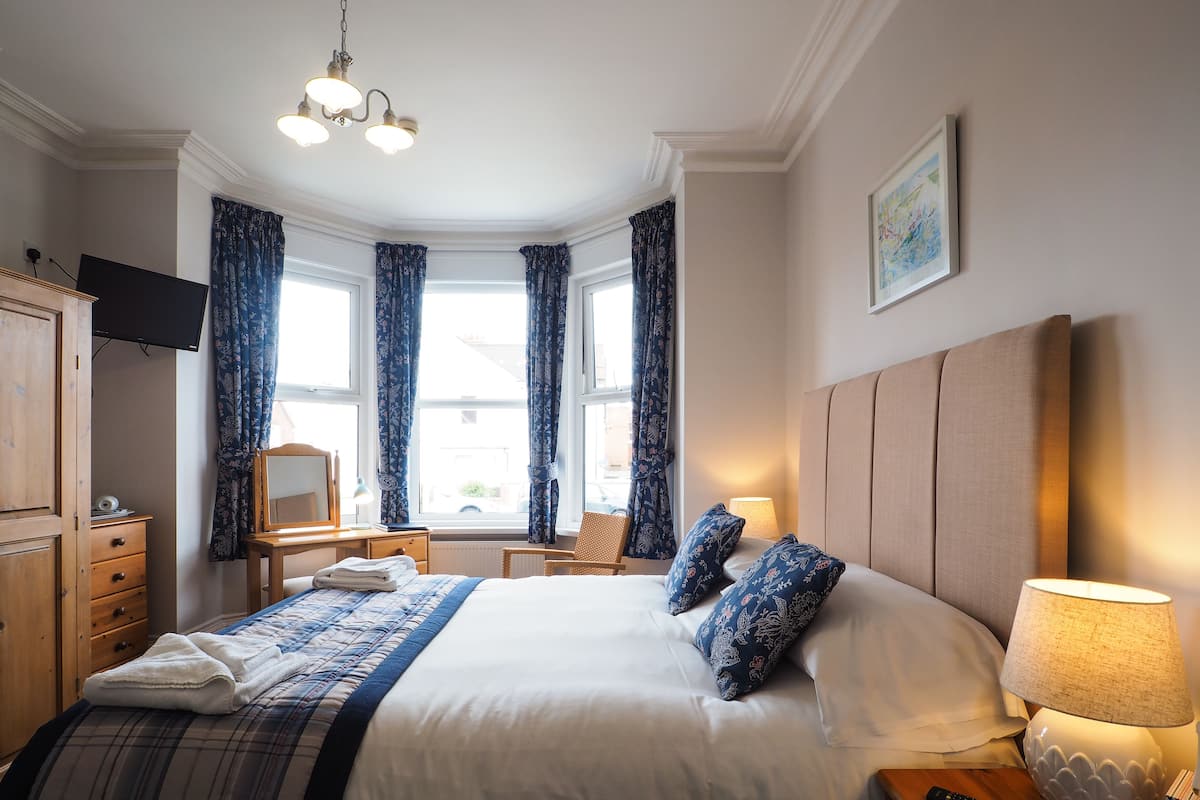 A hotel room with a large bed, a desk with a lamp, a chair, a television, and a window with curtains.