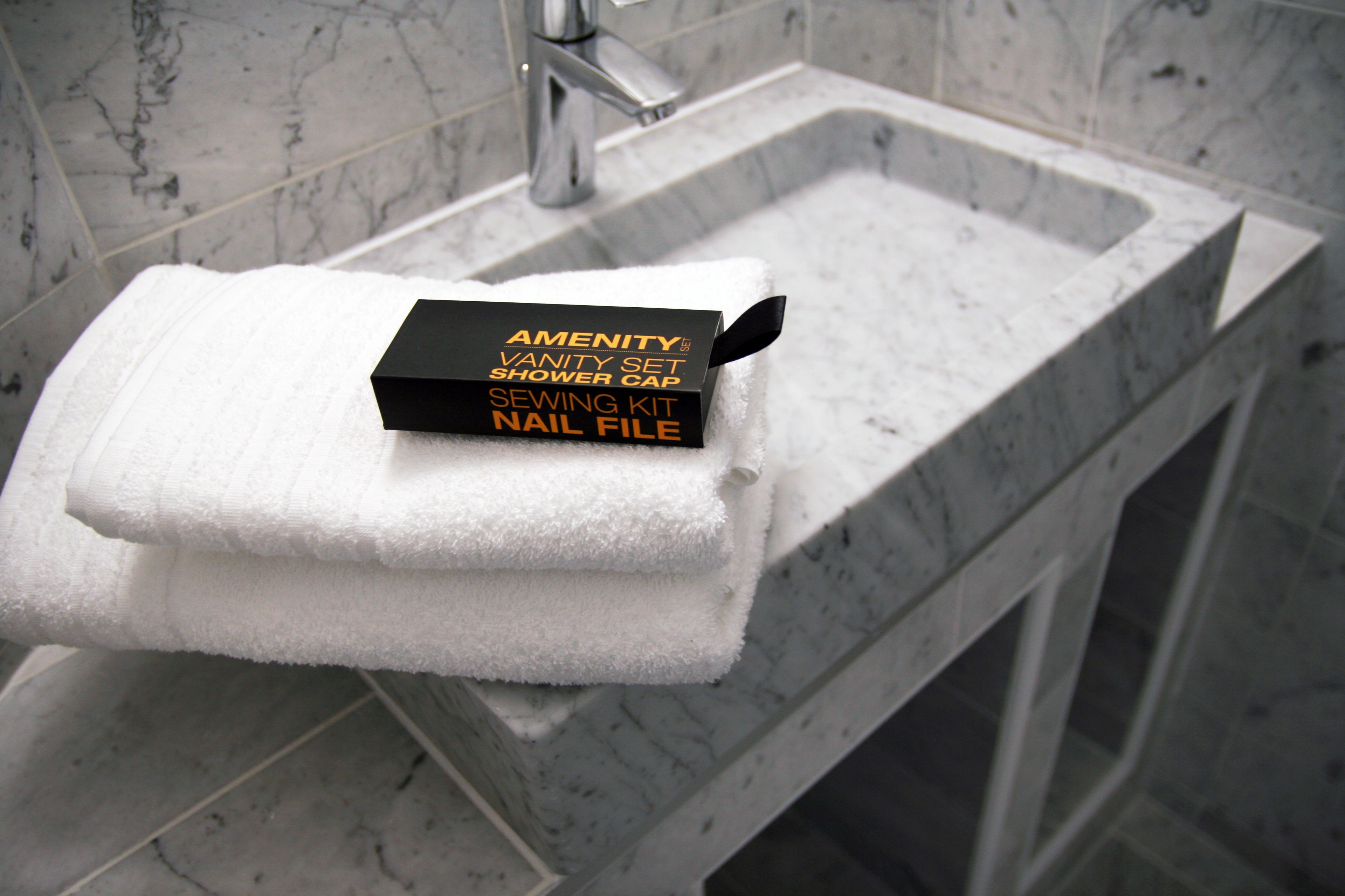 Une salle de bain avec un lavabo en marbre et une pile de serviettes posée dessus. Une boîte noire portant l’inscription « ENSEMBLE DE PRODUITS D’HÔTEL, CHARLOTE DE DOUCHE, KIT DE COUTURE, LIME À ONGLES » est placée sur les serviettes.