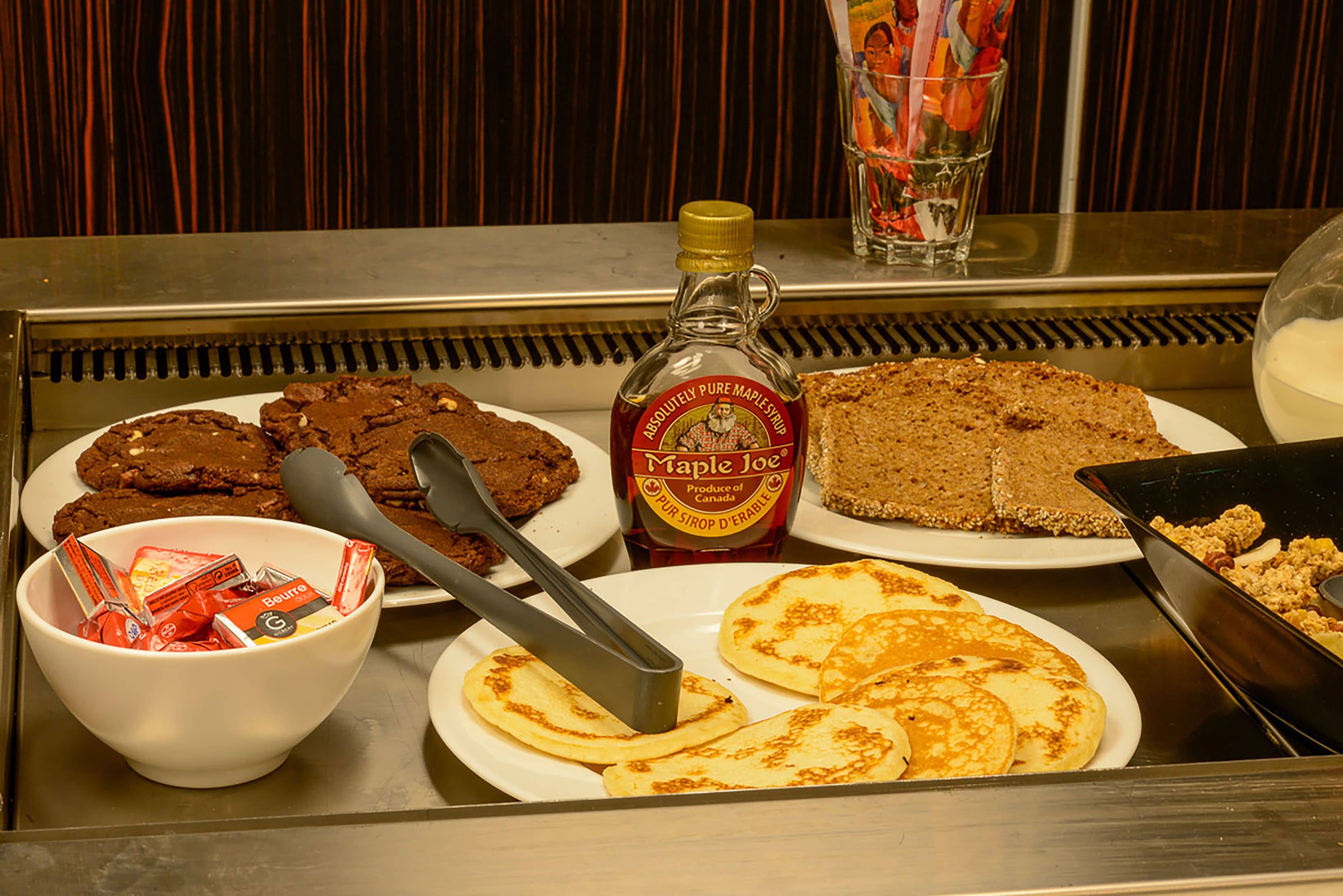 A breakfast buffet with plates of cookies, pancakes, and a bottle of maple syrup.