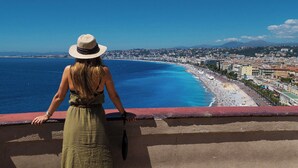 A woman in a green dress and straw hat overlooking a coastal city with a beach and buildings. - Hôtel & Appartements Monsigny (Nice)