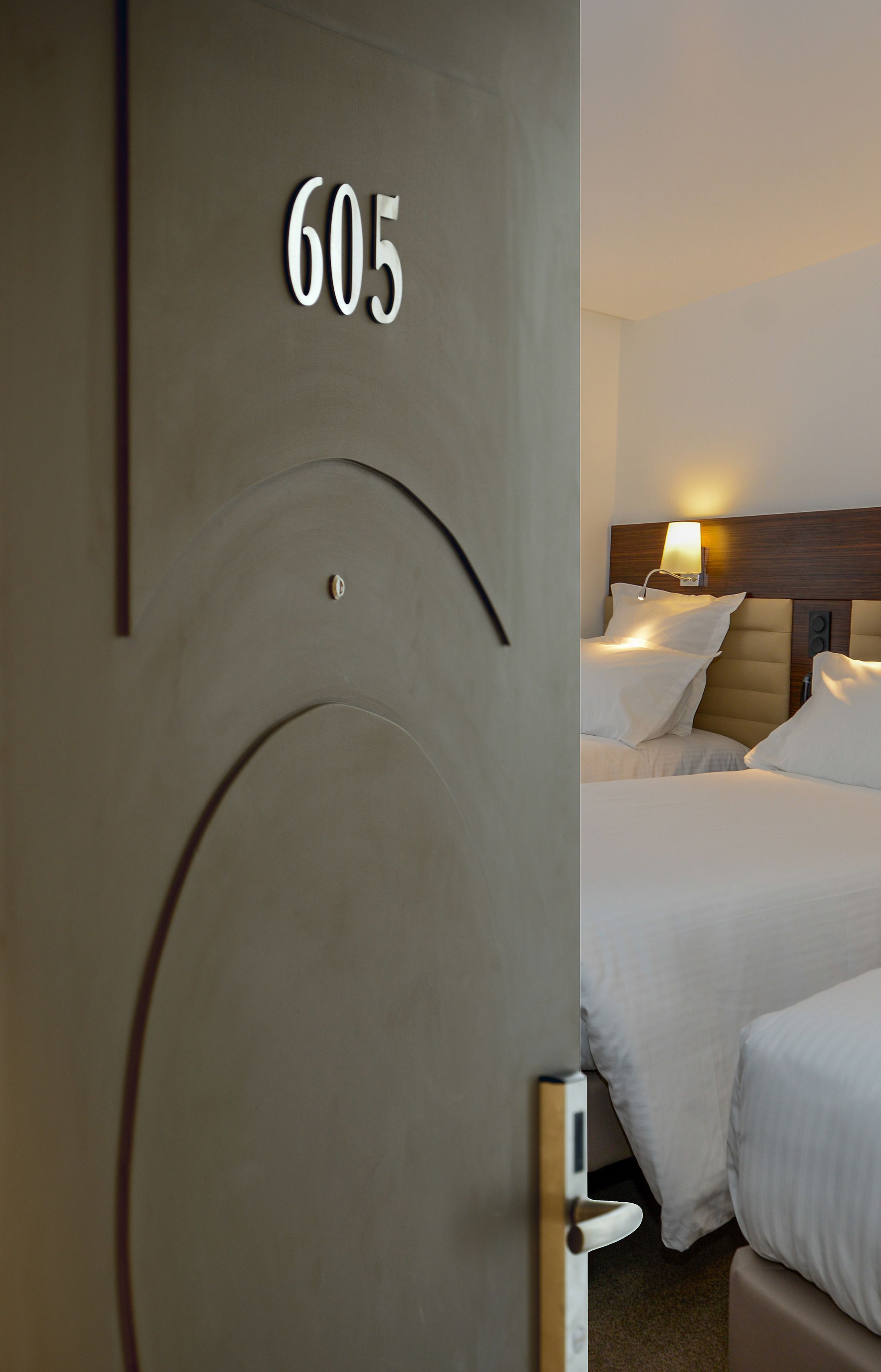 A hotel room with a door labeled "605," two beds with white bedding, a headboard with a lamp, and a wooden headboard.